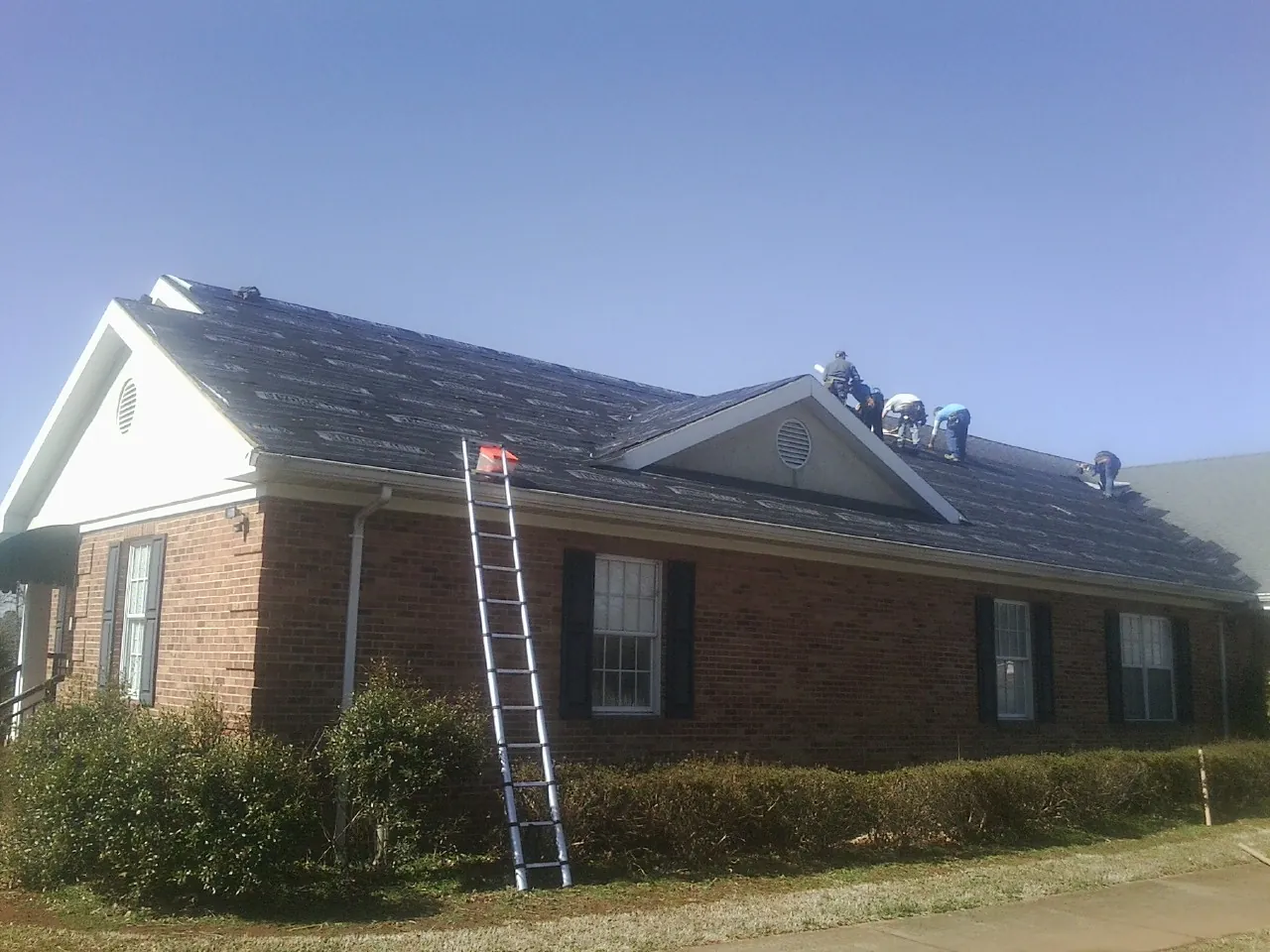 Crew tearing shingles off a brick ranch roof with a ladder set — Emergency Roof Repair work by our crew in Wimauma