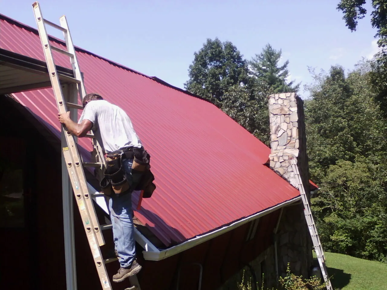Roofer on a ladder beside a red metal roof and stone chimney — Soffit & Fascia Repair by Roofing Brothers in Wimauma